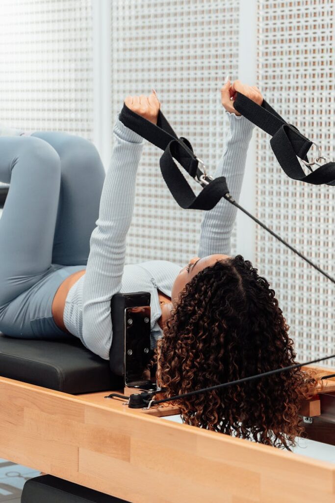 A woman wearing a gray tracksuit exercises on a Pilates reformer in a modern studio setting.